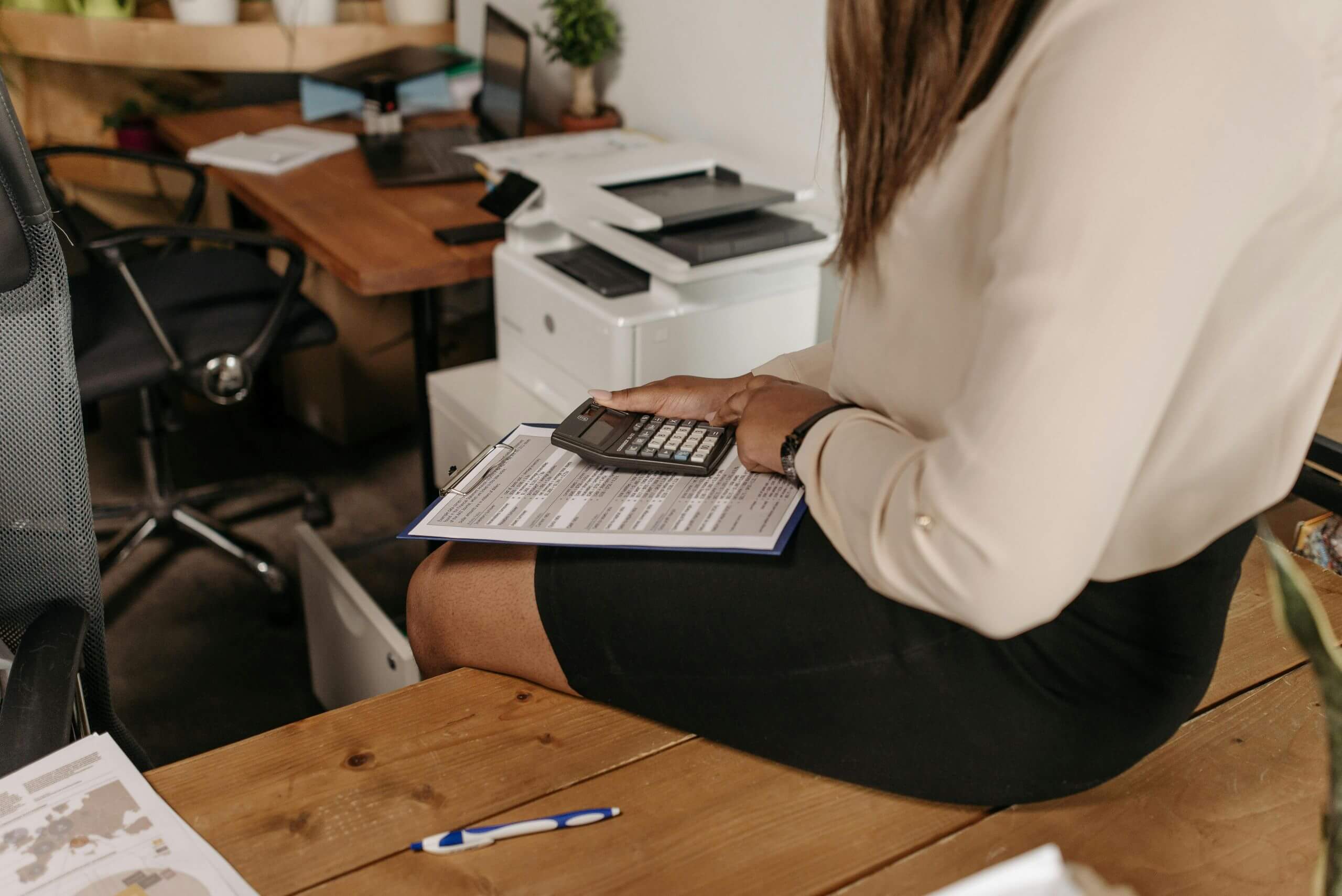 A woman in business attire sits on a desk, holding a clipboard and using a calculator. Papers, a pen, and office supplies are visible, highlighting a workspace environment during Cybersecurity Awareness Month.