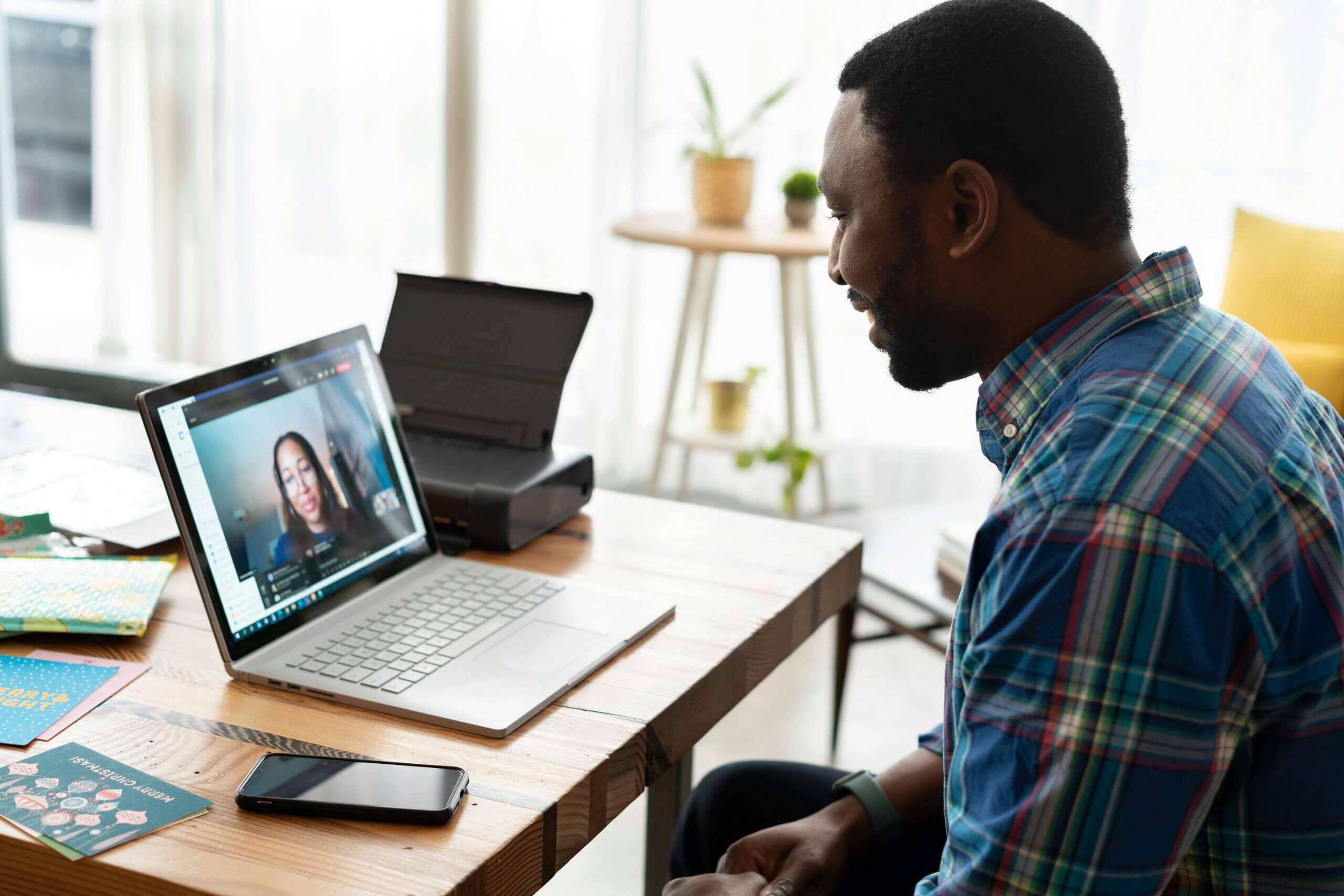 A man sits at a wooden desk, smiling during a video call on his laptop with a woman. The desk holds papers, a phone, and equipment for efficiency like a printer. A plant and sunlight brighten the background.