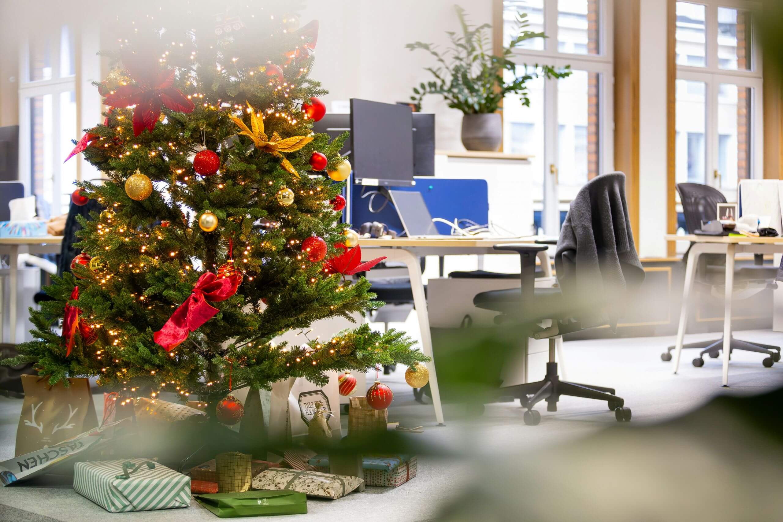A decorated Christmas tree with ornaments and lights stands in a bright, modern office with empty desks and chairs, showcasing a recent Office Equipment Refresh. Wrapped gifts are placed under the tree.