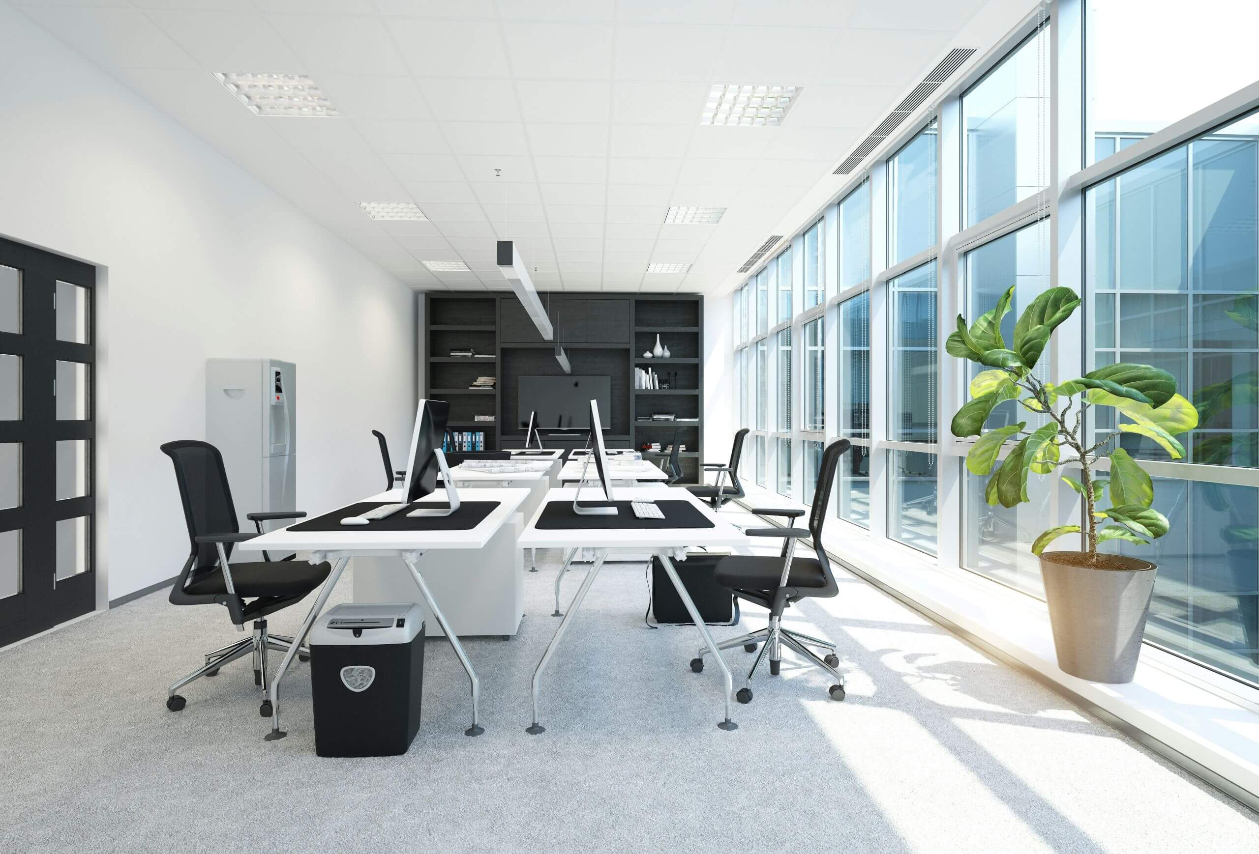 A modern, bright office with large windows, several black chairs at white desks equipped for a tech check with computers, a shelving unit in the back, and a tall potted plant near the windows.