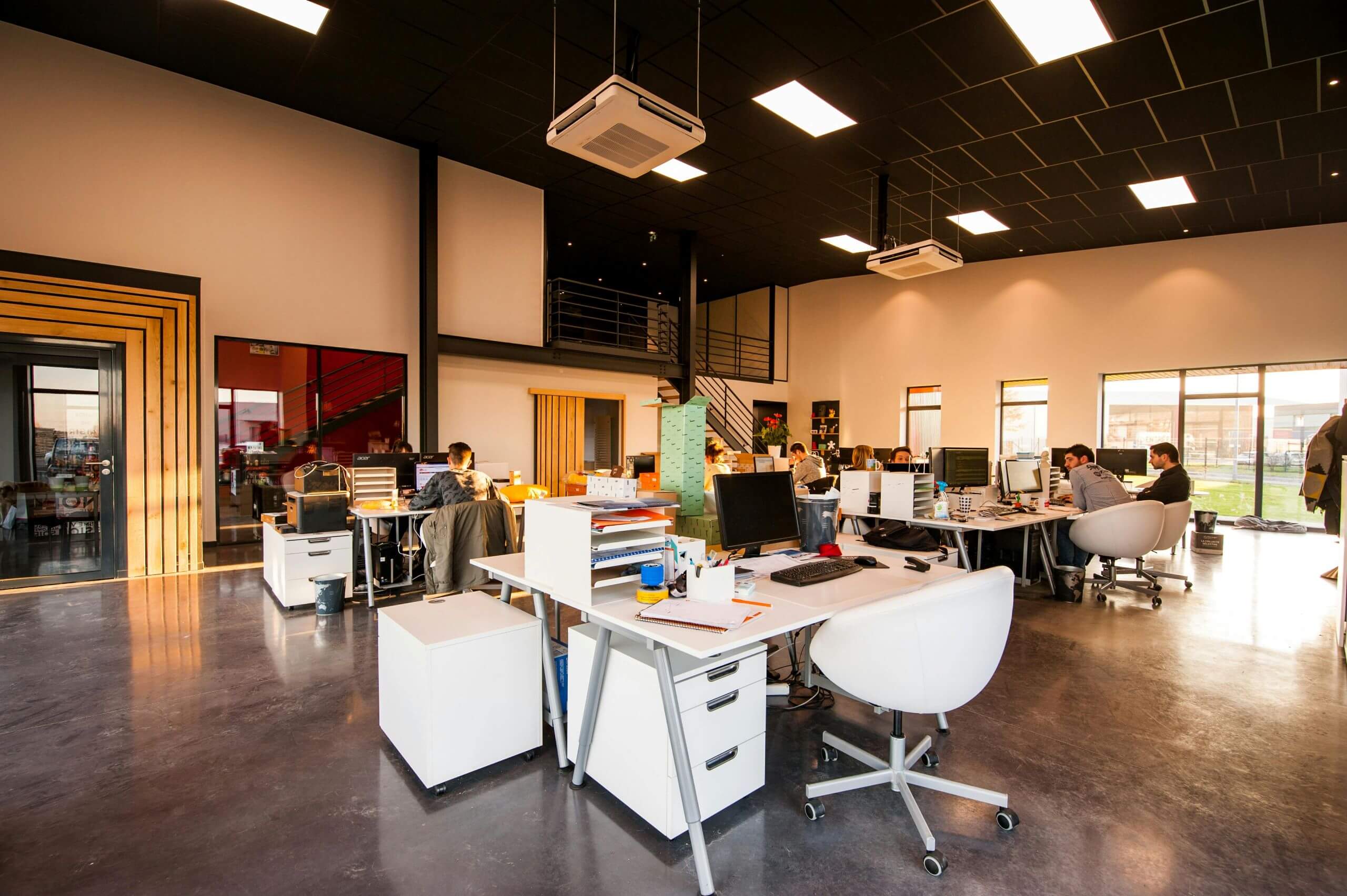 Modern open-plan office with several people working at desks, computers, and office chairs. Large windows let in natural light. The space has high ceilings, bright lighting, and contemporary decor, highlighting how essential document workflow solutions are