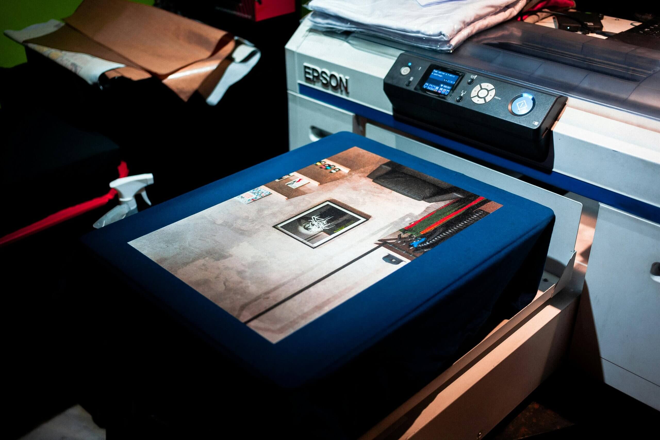 A close-up of Epson office equipment printing a colorful cat image in a picture frame on dark blue T-shirt fabric, with folded garments and printing supplies nearby.