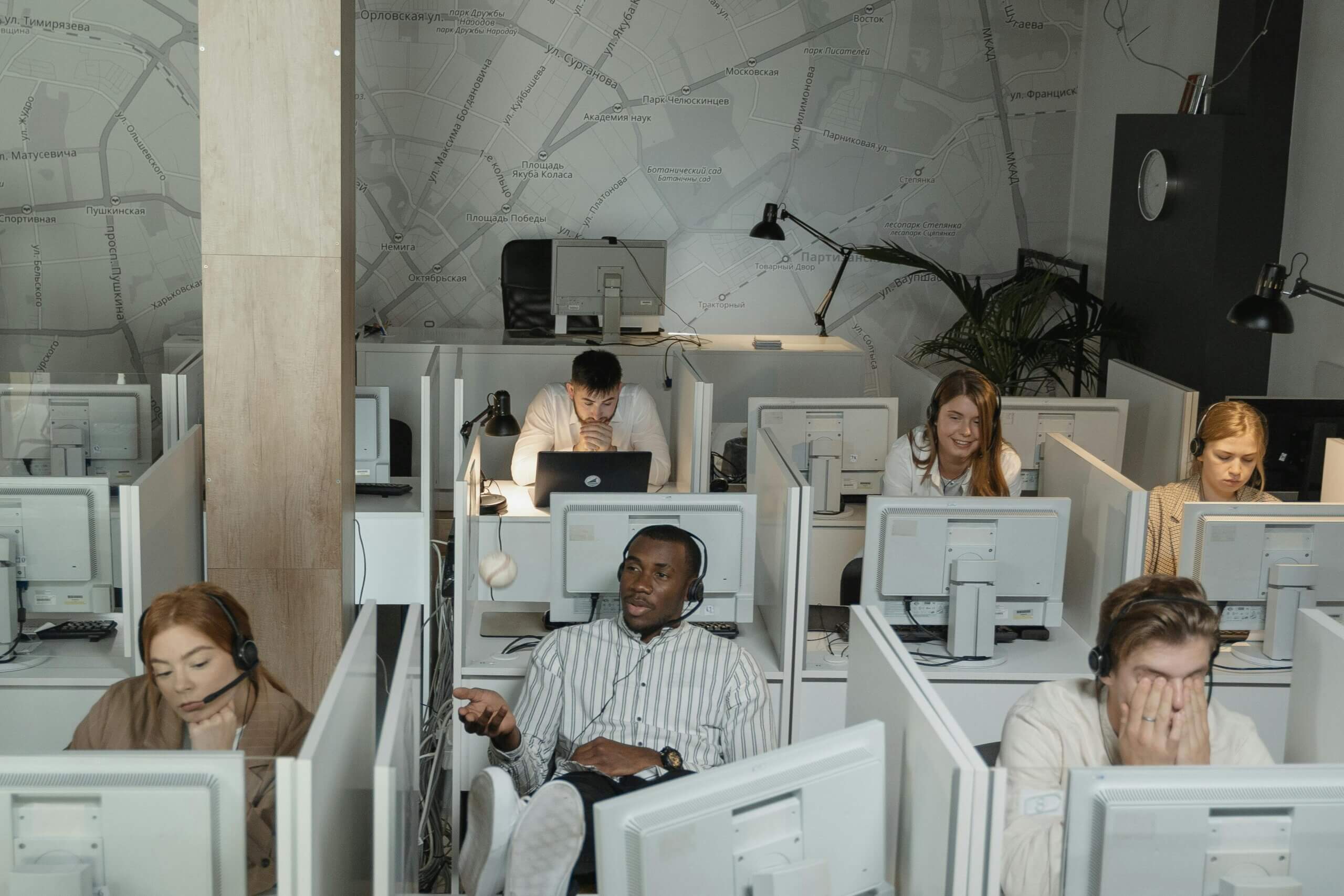 Six people sit in a modern call center, each at their own desk equipped with scalable office equipment and headsets. Some are talking or working, while one person covers their face, looking stressed. A city map decorates the wall behind them, highlighting modern office equipment upgrades in commercial workspace