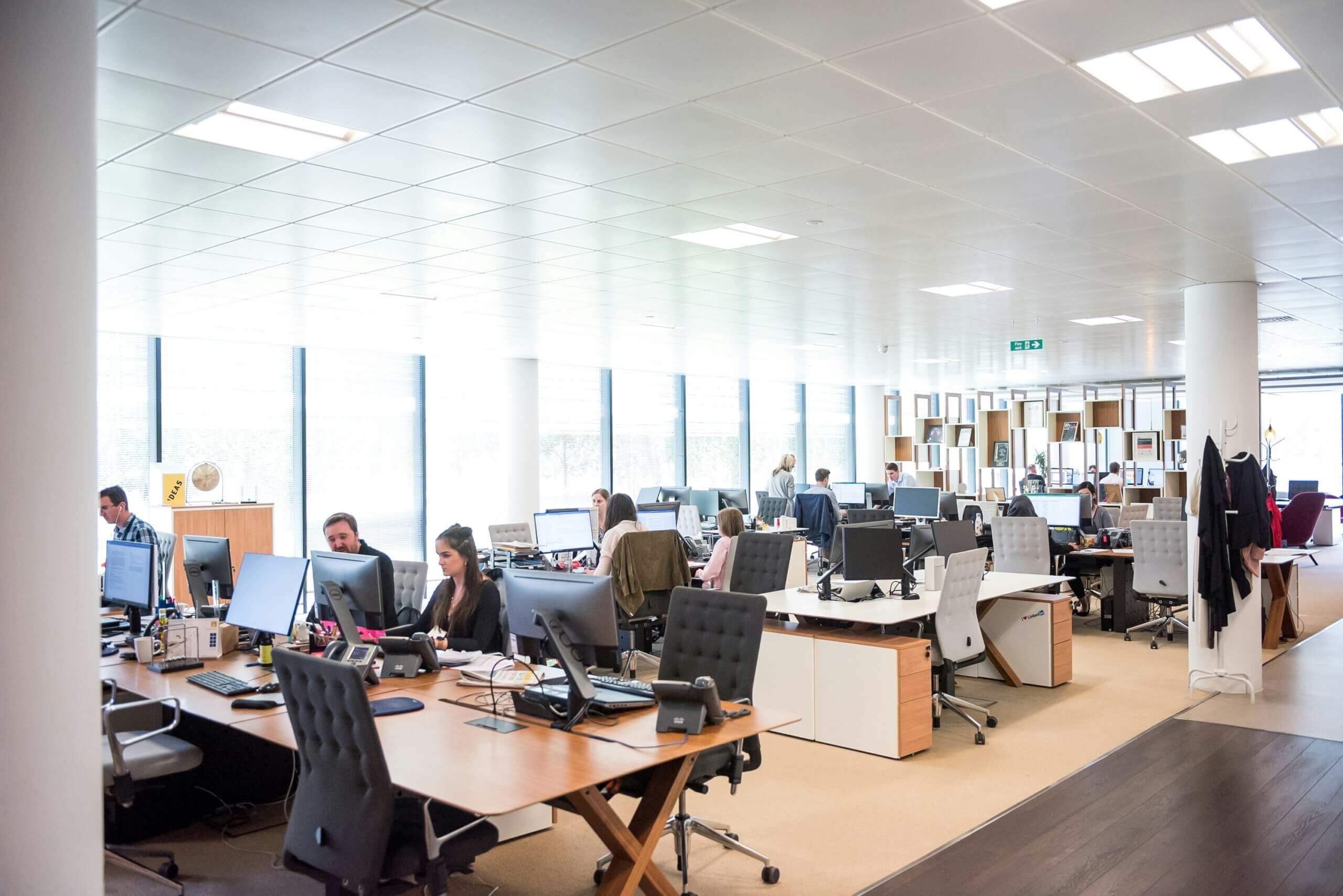 Modern open-plan office with several people working at desks with computers and equipment for productivity, large windows providing natural light, and shelves in the background. The space appears bright and organized.