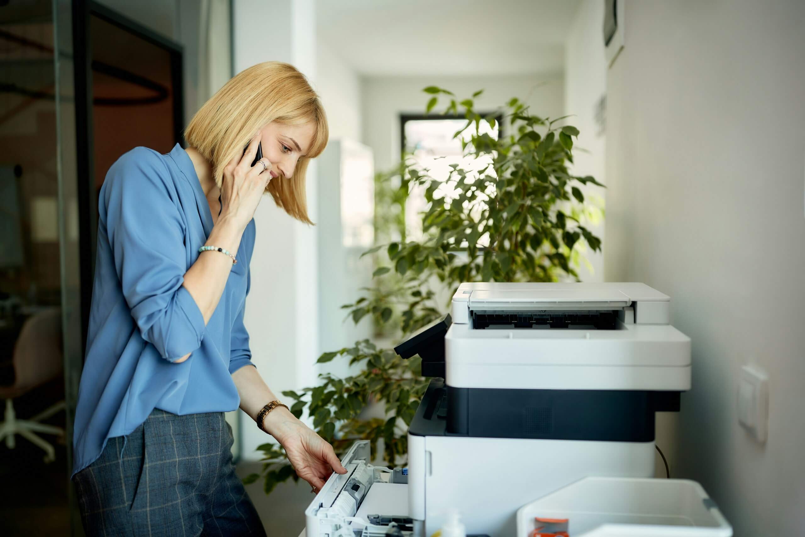 A woman with blonde hair wearing a blue blouse talks on the phone while using eco-friendly office equipment at a photocopier in a bright office with plants in the background