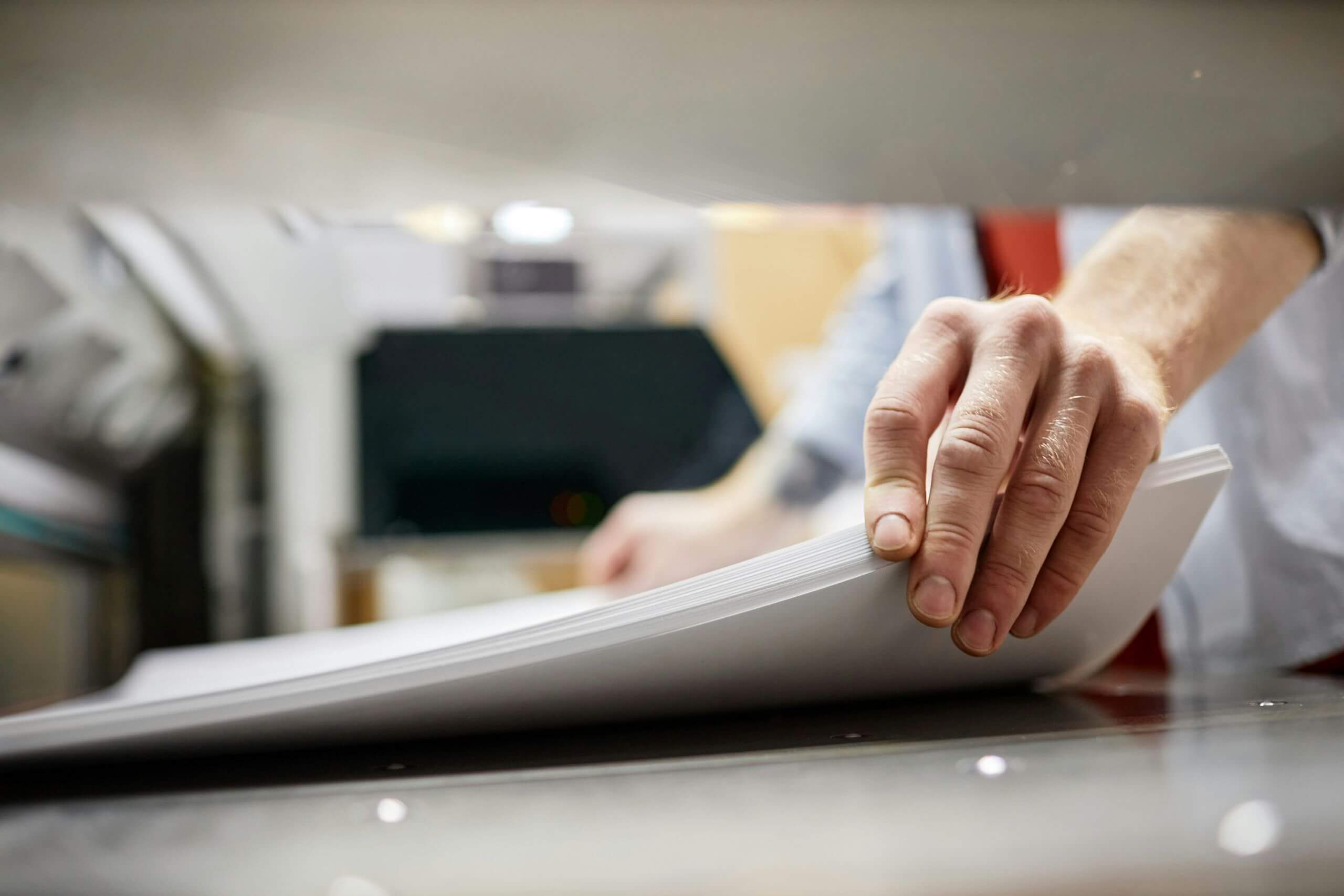 Close-up of a person's hand holding and aligning a stack of white papers on a cutting or printing machine like one of the high speed scanners, often found near high-speed scanners in an office or print shop setting.
