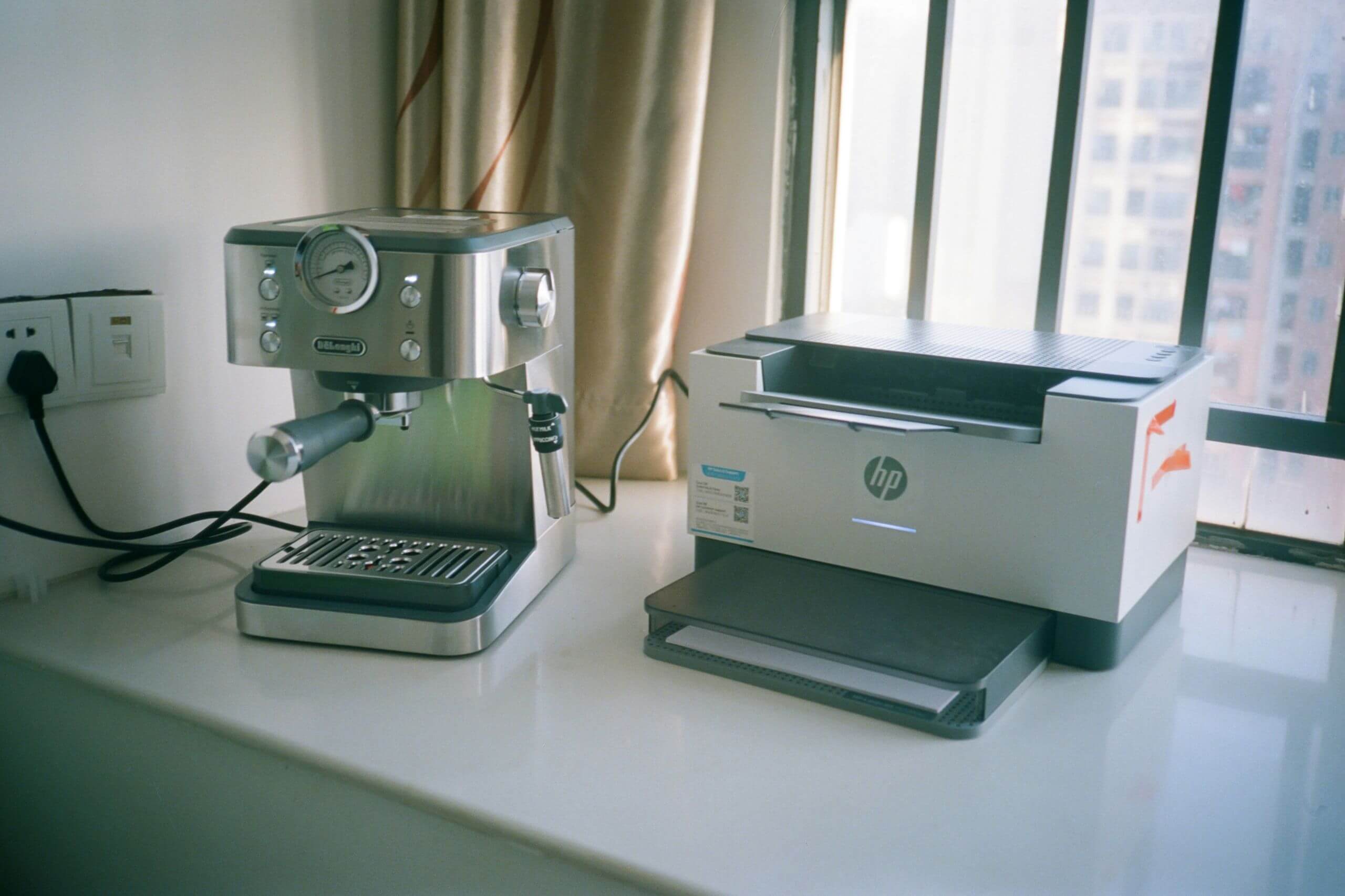 A DeLonghi espresso machine and an HP printer sit side-by-side on a white countertop by a window, all being shown from an office equipment company