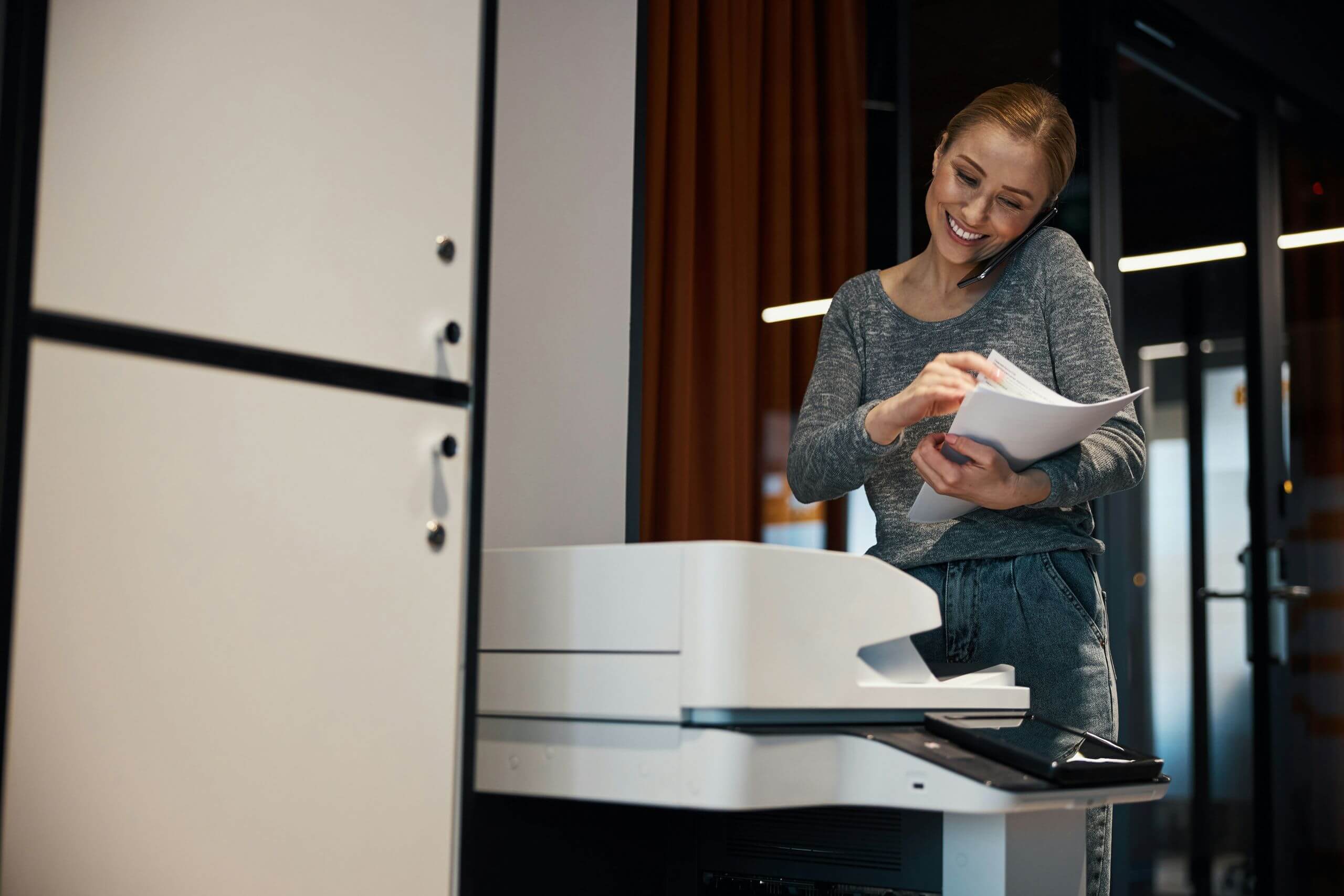 A woman smiles while talking on the phone and using a photocopier in an office, holding a stack of papers and multitasking, thanks to smart business printing solutions.