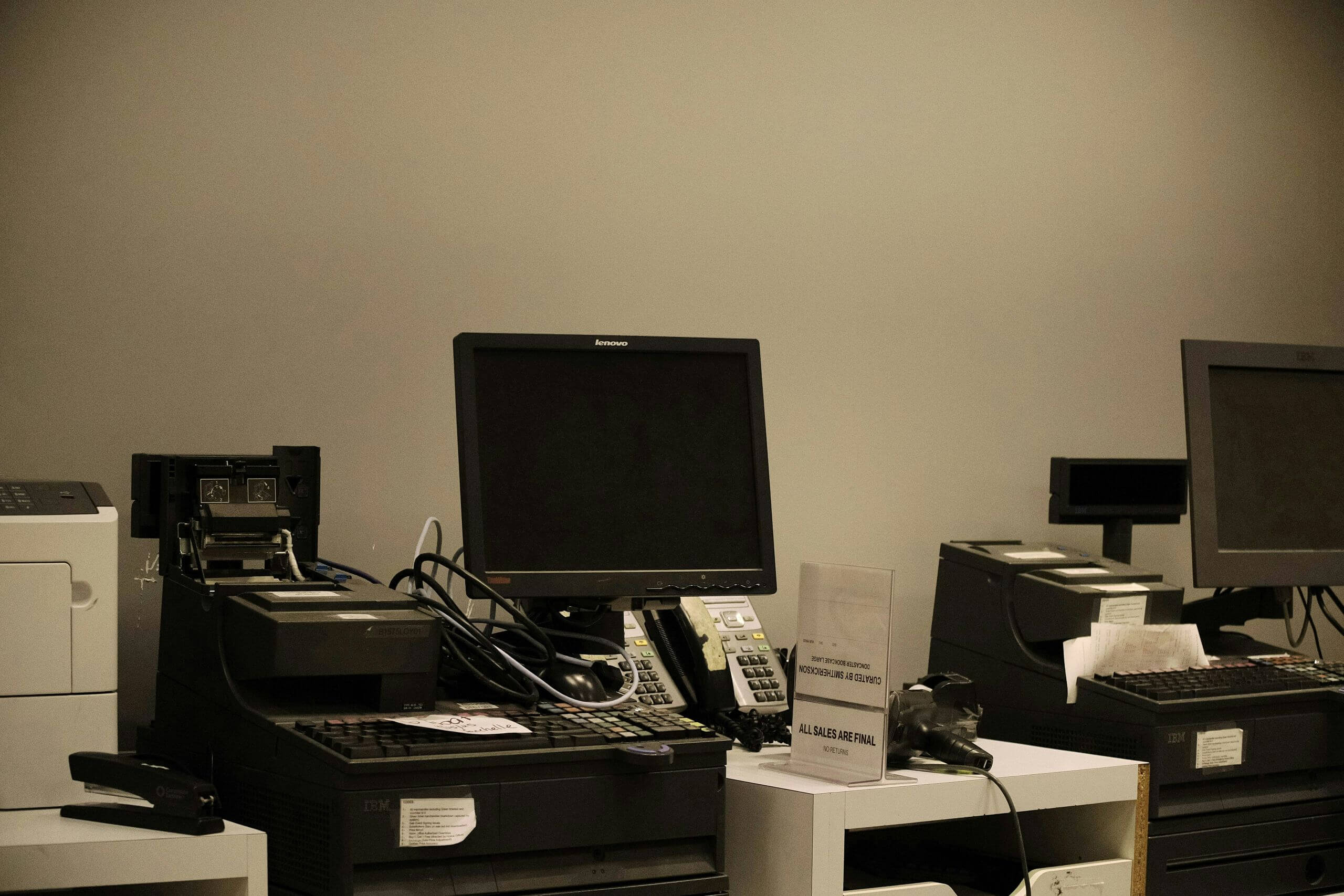 Two desktop computer workstations with black monitors, keyboards, and telephones sit on a white counter in a cloud-connected office setting. A printer, stapler, and various office supplies are also nearby.