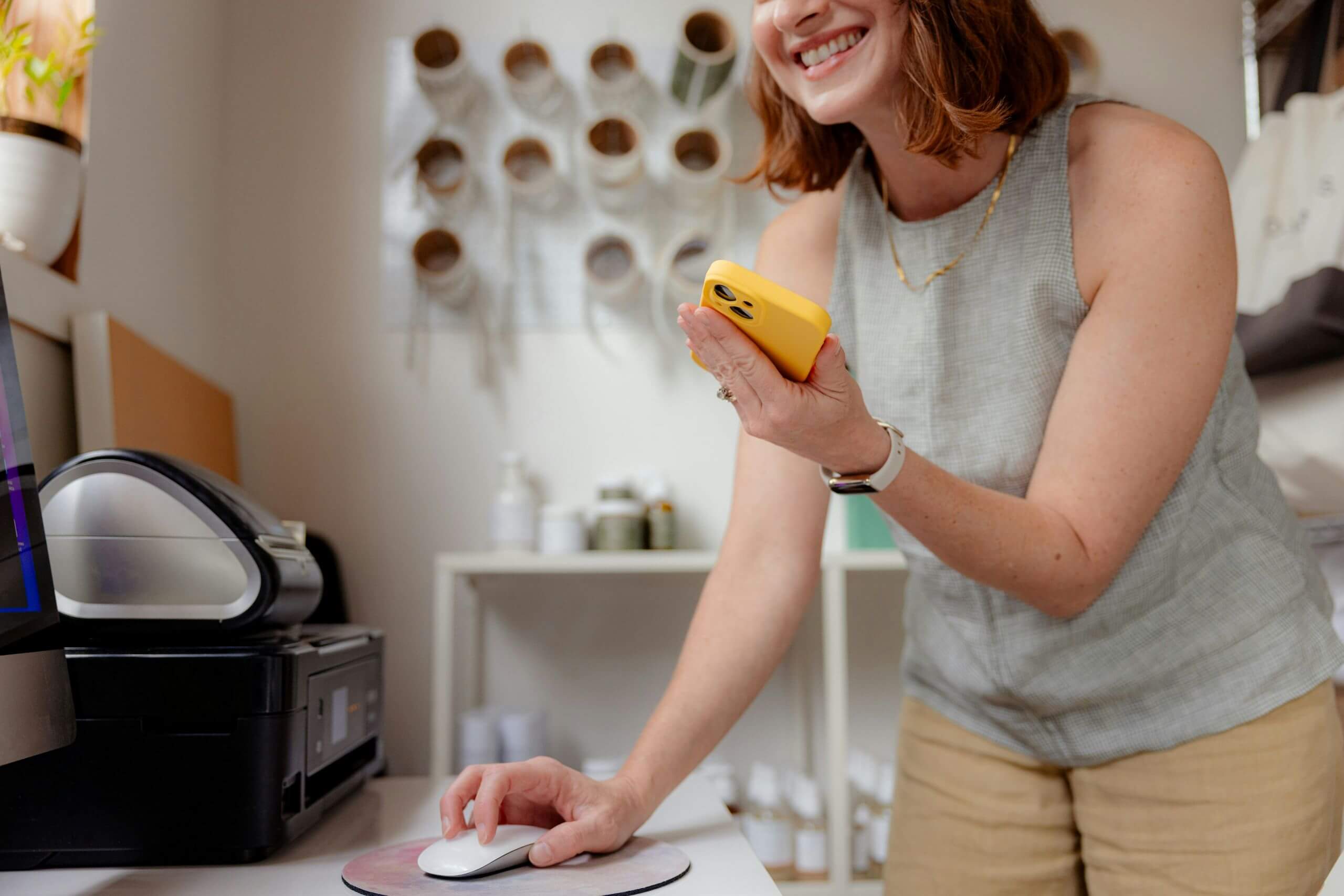 A woman smiles while holding a yellow smartphone and using a computer mouse in a home office or craft room, with shelves and supplies—perfect for exploring managed print services—in the background.