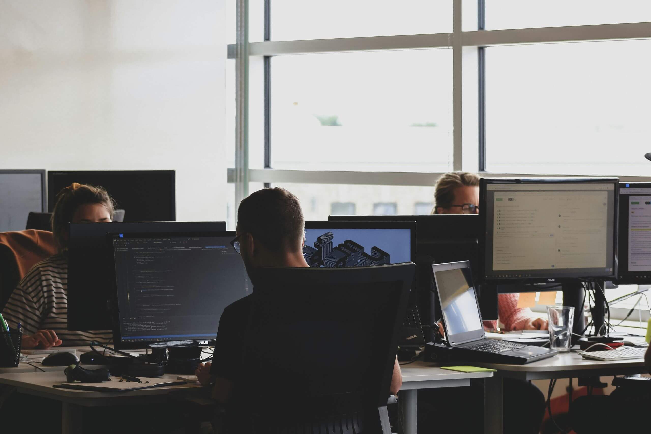 Three people work at computer desks in a modern office with large windows. Monitors, keyboards, and other office equipment are visible, creating a collaborative workspace atmosphere. Natural light fills the room, good for a business office technology