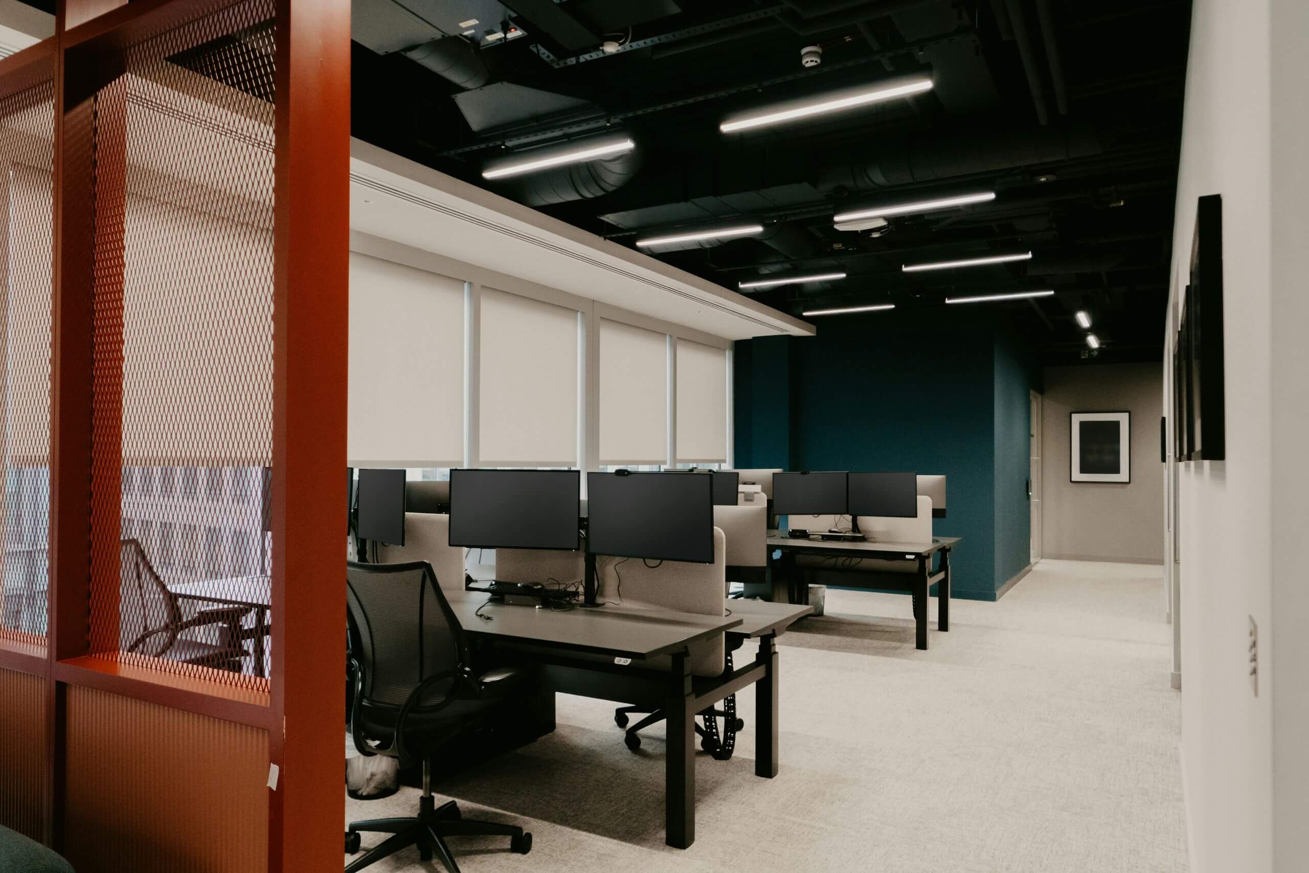 Modern office space for workplace productivity, shown with desks, multiple monitors, office chairs, and neutral decor. Ceiling lights illuminate the room, which has large windows covered with blinds and a blue accent wall in the background