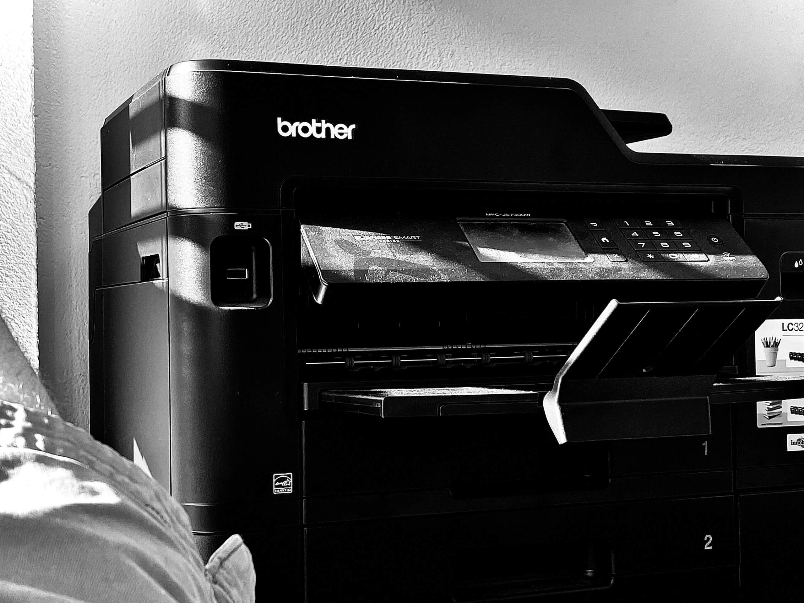 Black-and-white photo of a Brother brand printer near a wall, with an open paper tray and display panel visible, in a well-lit room, shown as one of the office technology solutions