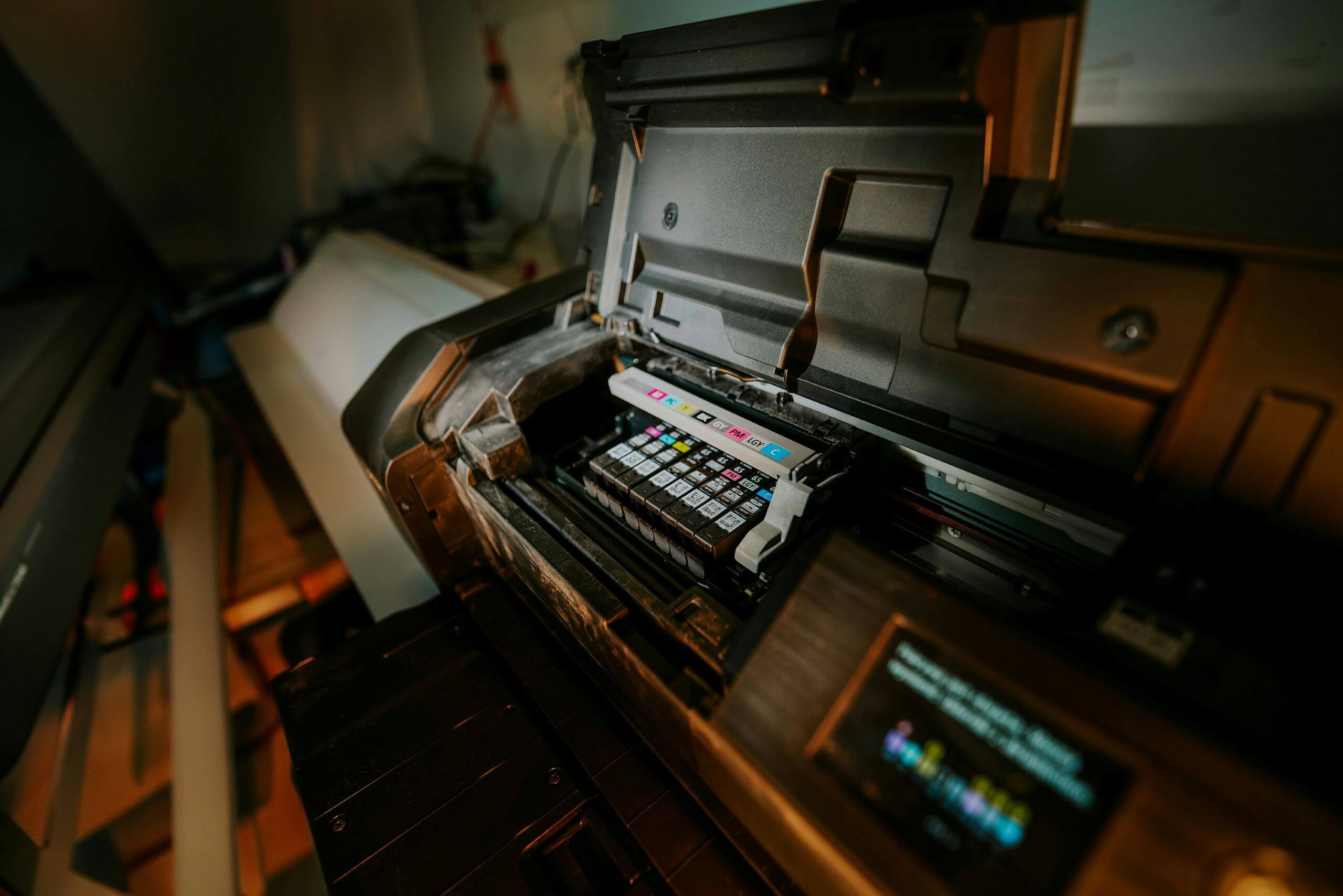 An open printer showing several installed ink cartridges inside, with a control panel displaying color information. The printer is situated in a dimly lit workspace showing the importance printer replacement for aging office equipment