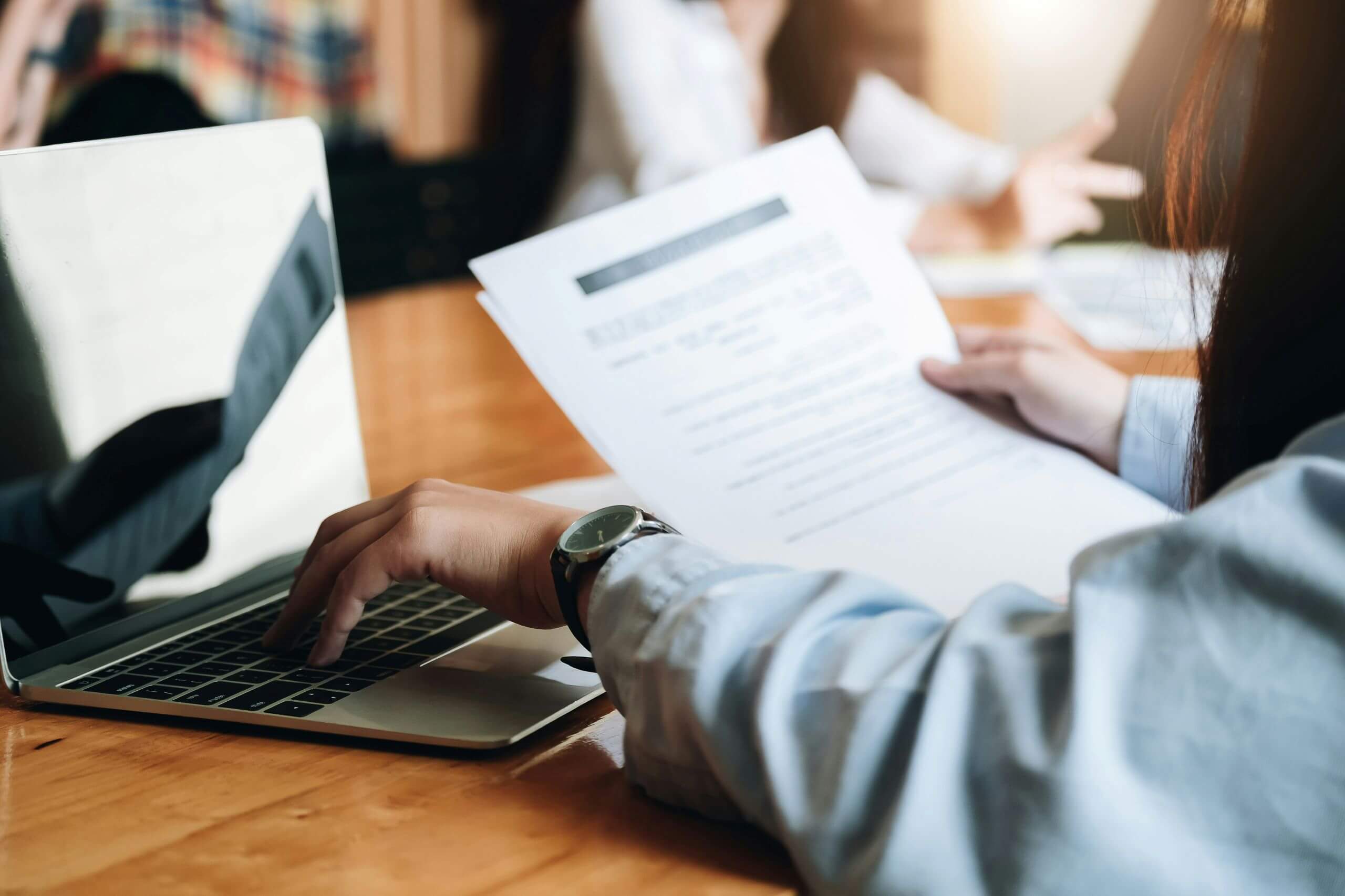 A person in a blue shirt holds documents in one hand and types on a laptop with the other, seated at a wooden table in a meeting room with blurred people in the background—perhaps reviewing their office printing budget while managing office printing costs during an important discussion.