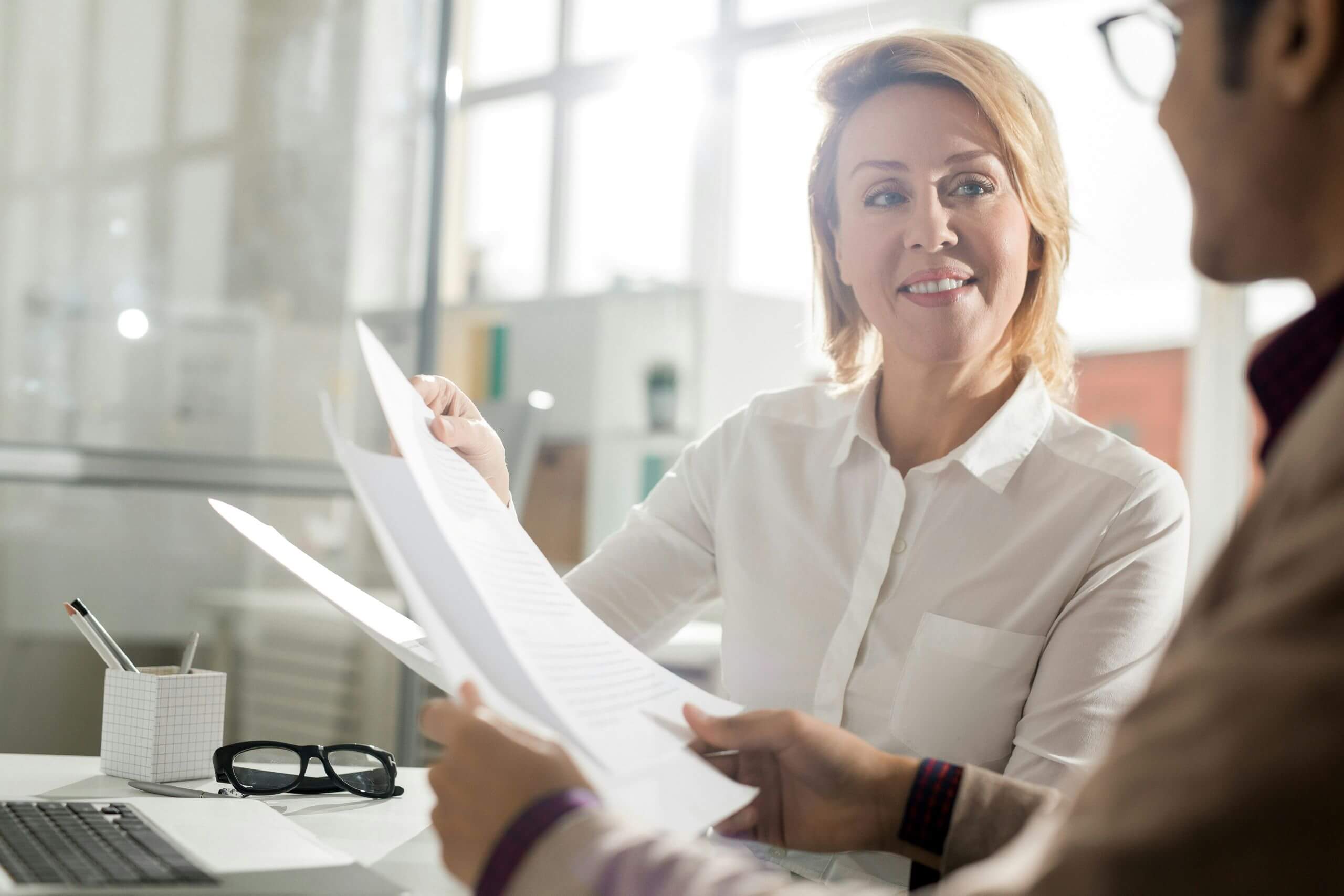 Two people sit at a desk in a bright office, exchanging documents. A woman in a white shirt smiles at a person across from her. A laptop, glasses, and cup with pens are on the desk.